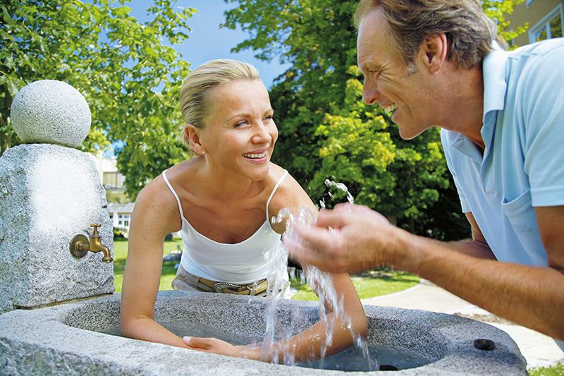 Ein fröhlicher Moment am Brunnen Ein Mann und eine Frau lachen und spielen mit Wasser an einem Brunnen im Freien.