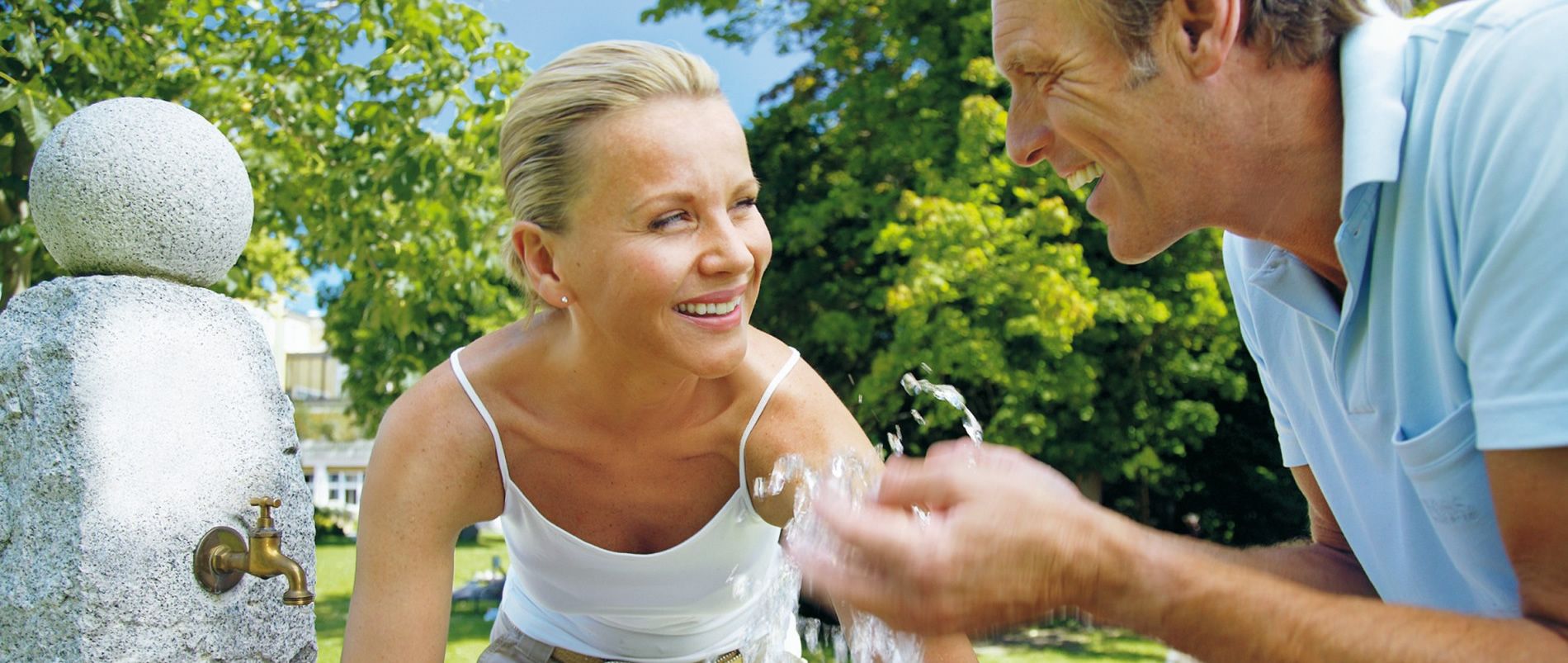 Spaß am Brunnen an einem sonnigen Tag Lächelnde Frau und Mann spielen mit Wasser am Brunnen an einem sonnigen Tag.